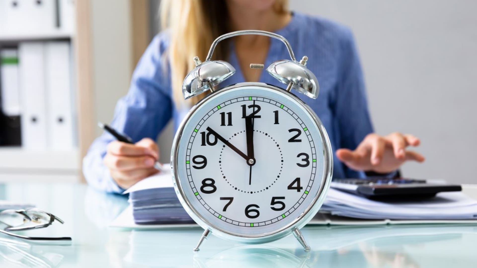 Reloj despertador plateado en primer plano marcando las 11:50, con una persona trabajando en segundo plano tomando notas y usando una calculadora.