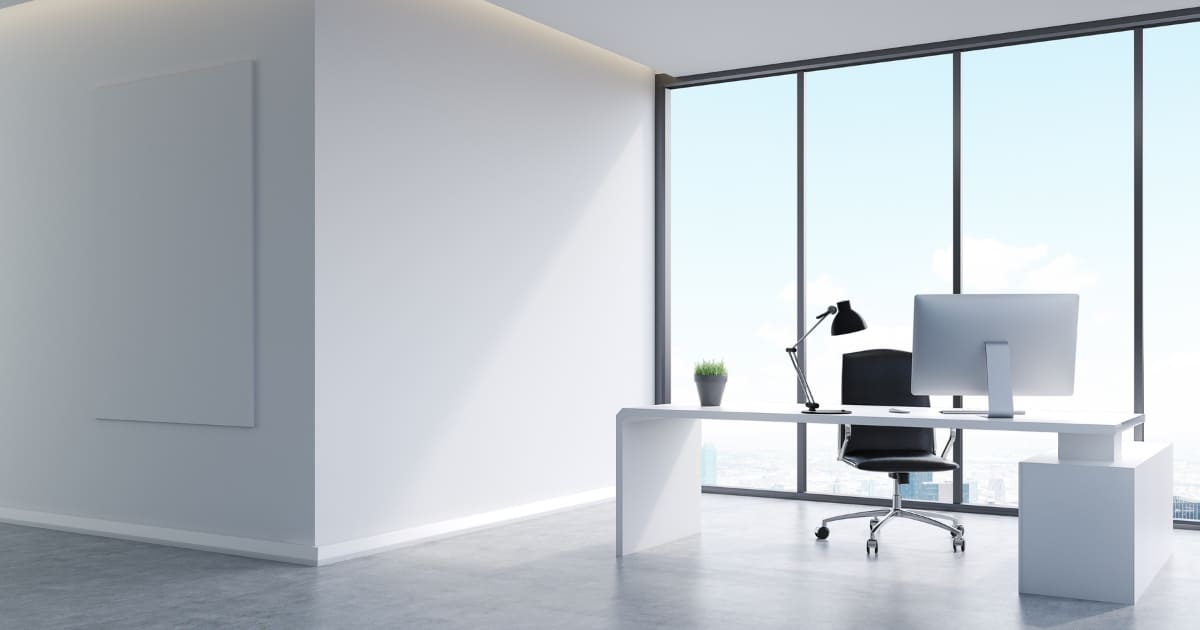 Espacio de oficina amplio y luminoso con escritorio blanco, ordenador, lámpara de escritorio y silla ergonómica frente a un gran ventanal con vistas a la ciudad; transmite modernidad, orden y productividad.