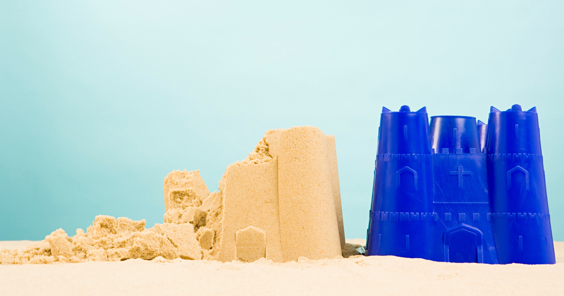 Castillo de arena parcialmente derrumbado junto a un molde de plástico azul en forma de castillo, sobre un fondo celeste.
