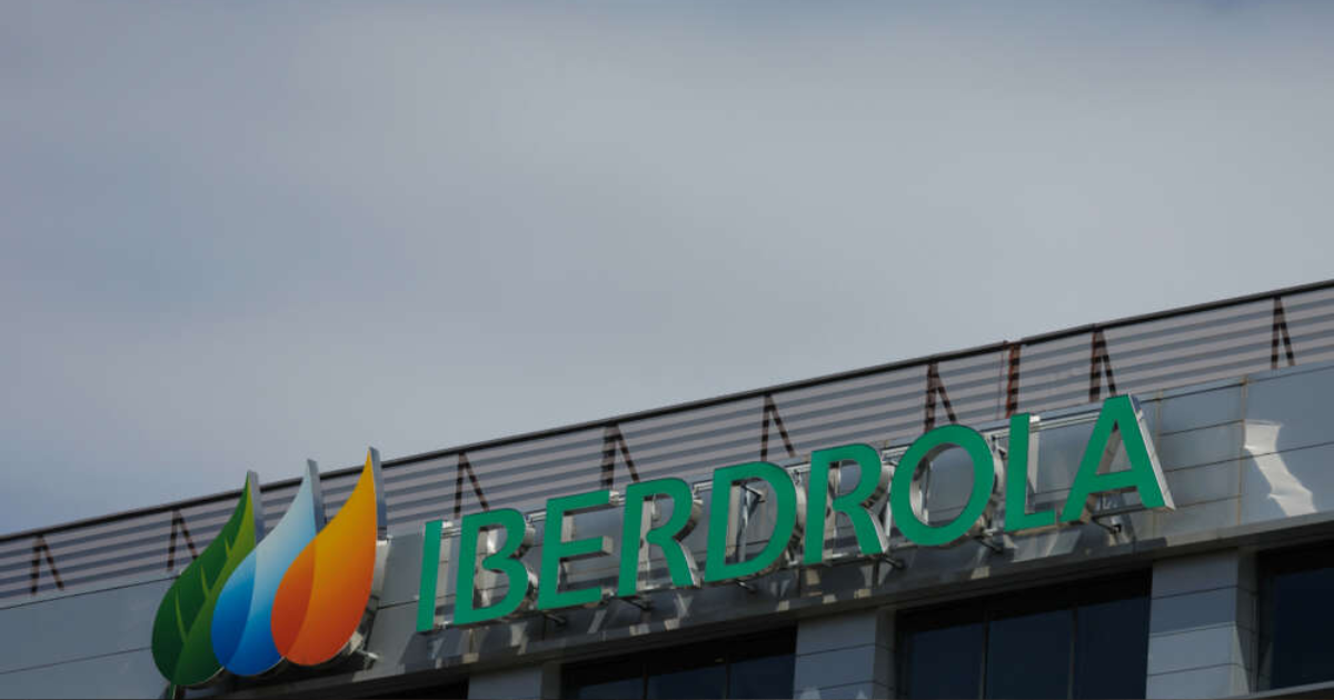 Fachada de un edificio con el logotipo de Iberdrola en color verde y su emblema de tres gotas multicolor, representando energía sostenible, sobre un cielo parcialmente nublado.